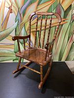 Front perspective view of the wooden children's rocker showing armrest and backrest details, with decorative painted background.