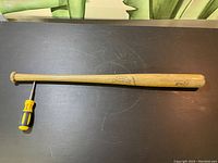 Full view of the vintage wooden baseball bat showing its length and shape on black surface with a screwdriver for size reference.