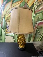 Front view of vintage table lamp showing ornate gold trimmed base on black pedestal with beige fabric lampshade with golden trim.