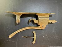 Top down photo showing the disassembled components of the vintage wall mounted corkscrew wine opener. The main lever arm assembly and the separate corkscrew with handle are displayed on a black surface.