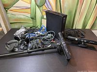 Black XBOX 360 console with Kinect sensors and various cables on a table.