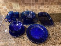 Overall view of all items arranged together on a kitchen countertop showing blue cobalt glass dishes including plates, bowls, mugs, platter and casserole dish.