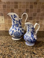 Front view of large and small blue and white ceramic pitchers showing floral and vine designs on a tiled counter.