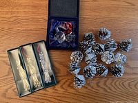 Photo showing three boxed pewter Santa Claus ornaments and silver painted pine cone ornaments spread out on a wooden surface with a blue velvet box containing the glass cat figurine.