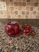 Cranberry glass apple and small flutter vase on granite countertop with tiled backsplash.