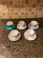 Five English fine bone china tea cups with matching saucers arranged on granite countertop showing floral and gold trim designs.