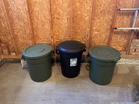 Overview of three garbage cans: one black with label in center, two green on sides with lids, placed against wooden wall.