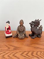 Three Japanese style decorative figurines displayed on a wooden surface against a white background: red ceramic Geisha, bronze-toned Samurai, and brass dragon.
