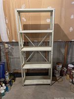 Front view of a white metal shelving unit showing five flat shelves and cross-bracing on the back, against a wall with paint cans nearby.