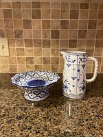 Porcelain footed plate and matching blue and white porcelain pitcher on granite countertop with tiled backsplash.