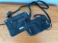 Two black crossbody purses on wooden surface, one larger purse with tags visible and smaller purse with buckle details