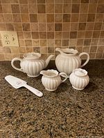 Full view of Grace's teaware white ceramic tea set including teapot, sugar bowl, creamer, and Mikasa cake server on granite countertop.