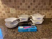 Overview of set showing 3 square dishes stacked, 2 round pots with spouts stacked, 3 detachable black handles, one in original box, and a cradle stand.