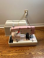 White and pink portable sewing machine placed on base with attached foot pedal, accompanied by a white plastic hard carrying case behind it.