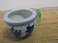 Side perspective of the blue and white ceramic flower pot placed on carpet, next to a green plastic bottle for scale.