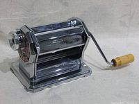 Photo showing side profile of shiny chrome metal pasta maker with crank handle made of wood on the right side, resting on cloth background.