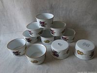 Set of 10 white ceramic ramekins with fruit decorations and gold rims arranged together.