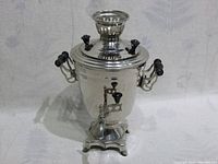 Front view of the polished silver-tone vintage Russian samovar on four-legged decorative base with black wooden handles and spigot.