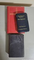 Three vintage Bibles stacked on a grey surface: a red hardcover Bible, a black cloth-covered Bible, and a compact navy blue Bible with gold text titled 'New American Standard New Testament Old Testament Prophecy Edition'.