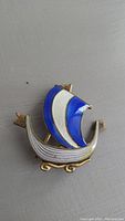 Front view of antique enamel brooch shaped like a Viking ship, showing blue and white sail and gold tone metal with decorative details.