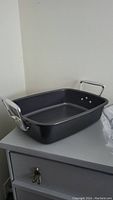 Photo of the black rectangular roasting pan with silver metal handles on a grey surface against a white wall.