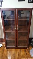 Front view of wooden display bookshelf cabinet with two glass doors and brass handles, showing the shelves inside.