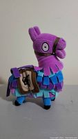 Side angle view of the purple and blue Fortnite llama stuffed animal with brown saddlebag and cartoon eyes.