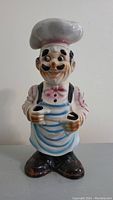Front view of ceramic chef figurine with utensil holes in hands. White hat, blue apron, and detailed painted features.