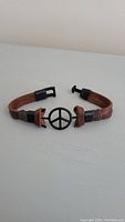 Front view of brown leather bracelet with black metal peace symbol and clasp visible.