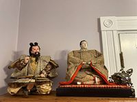 Two seated Asian figures and an ornate metal decorative item on a flat surface against a light wall
