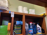 Wide shelf view showing multiple cleaning supplies including Windex, Lysol, Swiffer wet cloth boxes, and cleaning sponges with paper towels and napkins above.