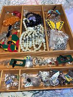 Wooden tray with divided sections containing an assortment of costume jewelry including necklaces, pins, and earrings.