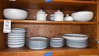 Scooped view of white Dansk Bistro dinnerware including stacked plates, bowls, and covered serving pieces with blue rim detail inside a wooden cabinet.