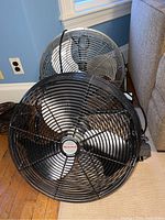 Two Honeywell commercial grade fans with black metal grilles and silver blades positioned on the floor near a blue wall and couch.