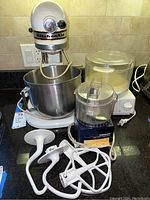 KitchenAid cream-colored stand mixer with a stainless steel bowl and three white attachments (dough hook, flat beater, whisk) positioned next to Cuisinart food processor with clear container and white base on a black kitchen counter