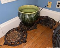 Green ceramic planter with floral relief pattern and visible cracks on the exterior, sitting on wooden floor near wall outlet.
