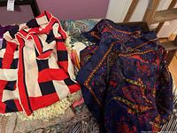 Scarves piled together showing a red, cream, navy geometric striped scarf with fringed edges and a dark paisley patterned scarf
