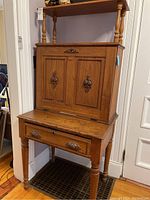 Photo showing the front of the wooden secretary desk with closed fold-down door and drawer below, featuring decorative metal hardware and turned legs.