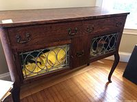 Front view showing three drawers and two leaded glass doors with metal handles, with visible yellow items inside cabinet behind glass.