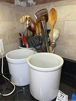 Photo showing two white ceramic crocks on a kitchen counter filled with various cooking utensils.