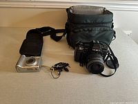 Two cameras on a surface includes a silver point-and-shoot camera in black carrying case and a Minolta Maxxum 7000 SLR camera with strap, earbuds, and large gray carrying bag behind