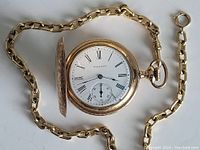 Watch open revealing white enamel dial with Roman numerals, subsidiary seconds dial and attached gold-tone chain