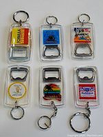 Overview of six vintage bottle opener keychains with various branded and scenic inserts under clear plastic cases. Includes Cowichan, Alberta, Budweiser, and tropical motifs.