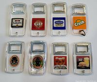 Eight vintage bottle openers arranged in a grid, each with a clear plastic body with a metal opener at the top. Each has a circular colorful label featuring various brewery and soda brands.