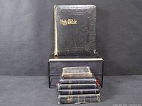 Six antique Holy Bibles stacked with one standing upright showing worn black cover with gold embossed title