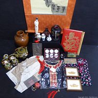 Photo showing a collection of Asian art and collectibles including pottery, textiles, figurines, framed art, and a decorative book arranged together.
