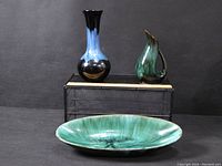 Photo showing the three Blue Mountain Pottery pieces: a blue and black vase, a green pitcher, and a large green bowl, arranged on a black surface against a black background.