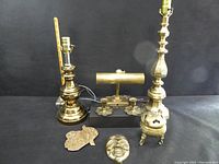 Wide view showing two large brass candlesticks, brass desk lamp, brass flower candle holders on black base, brass face mask, brass incense burner, and brass trivet.