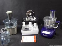 Wide view of silver juicer on the left, white attachments container with discs and white items in the center, and blue KitchenAid food processor on the right with user manual in front.