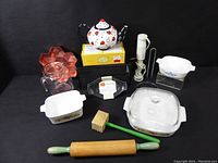 Photo of assorted kitchenware items laid on black surface showing ceramic teapot, glass pitcher, wooden rolling pin, wooden tenderizer mallet, plastic kitchen utensils, baking dishes, and star-shaped trays.